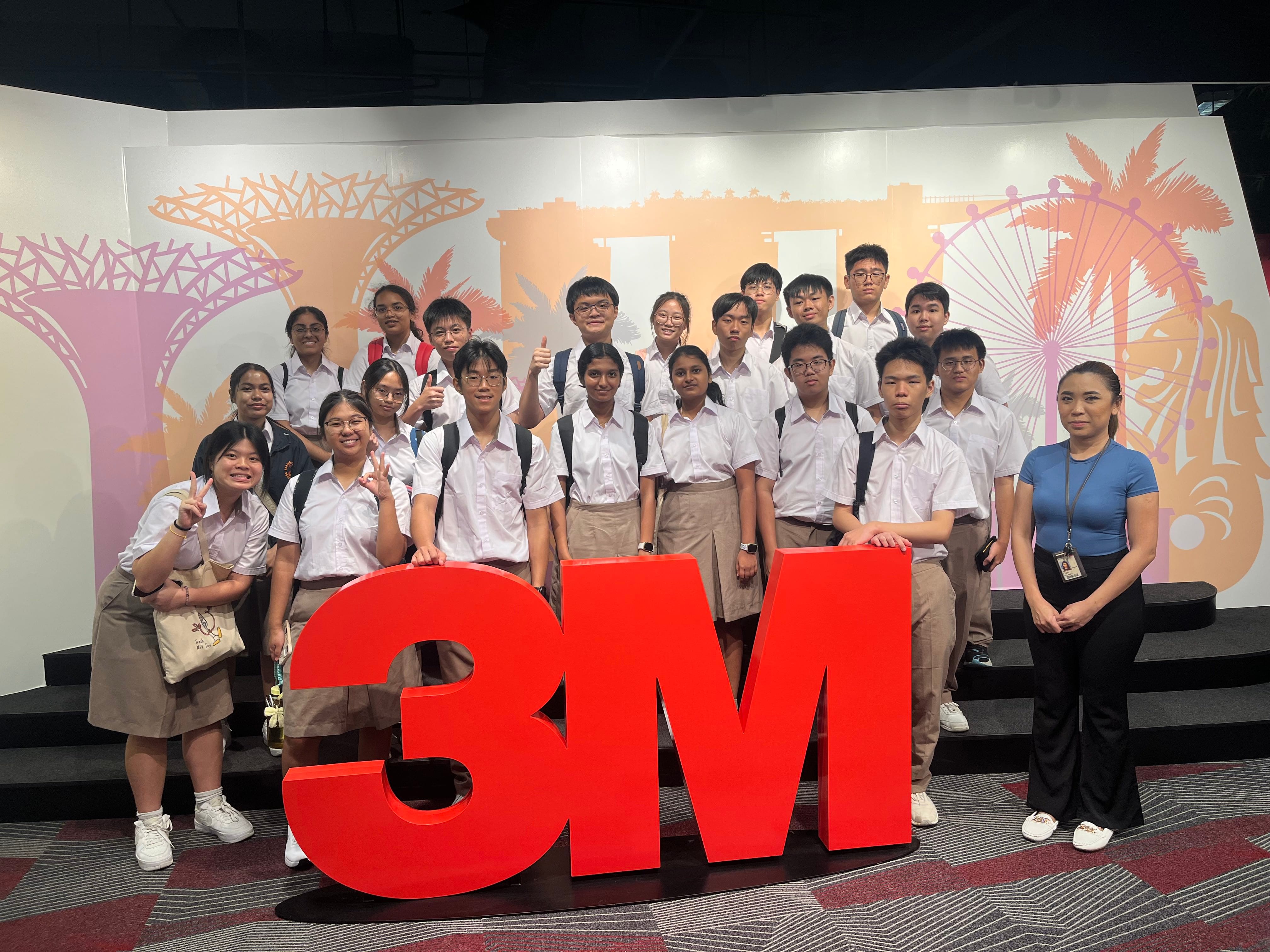 A large group of students in school uniforms poses in front of a colourful Singapore-themed mural, gathered around a large red 3M logo display